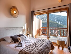 Een slaapkamer met een groot bed, uitzicht op een balkon met stoelen en een berglandschap.