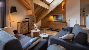 Een loft-achtige woonkamer met een schuine plafond, houten balken, een aan de muur bevestigde televisie en een zithoek.
