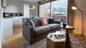 Een moderne woonkamer met een grijze bank, een bijzettafeltje met boeken en een kopje, en een groot raam met uitzicht op bomen.