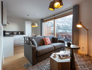 A modern living room with a grey sofa, a coffee table with books and a cup, and a large window with a view of trees. - Apartment in Vaujany Near Alpe D'huez Slopes (Vaujany)