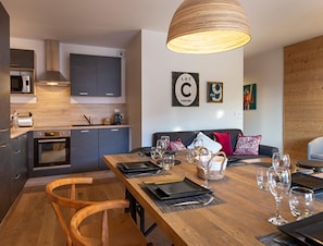 Une cuisine moderne avec des meubles gris, une table à manger en bois pour quatre personnes et un grand luminaire suspendu.