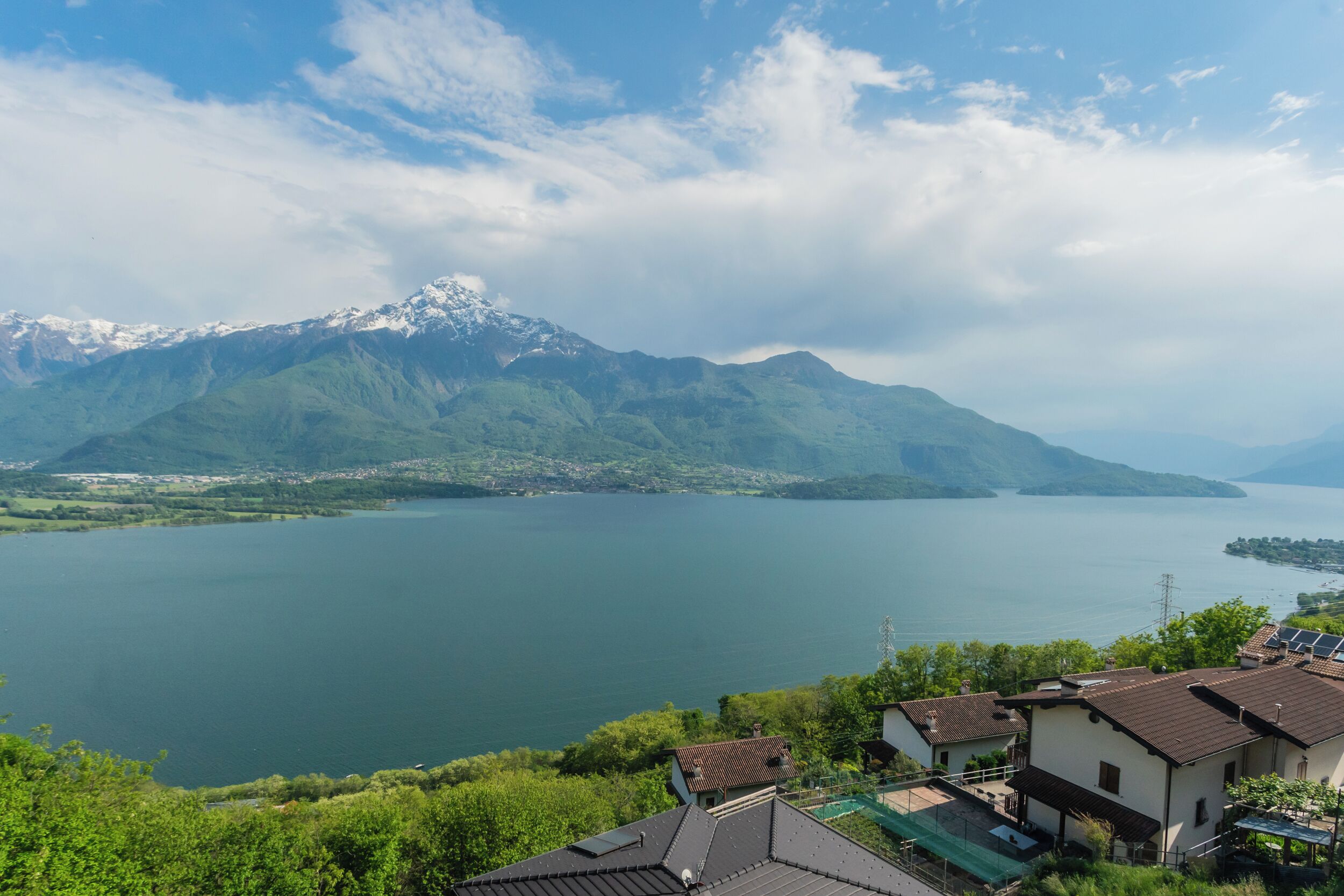 Vista di un lago con montagne sullo sfondo, case con tetti spioventi e cielo sereno.