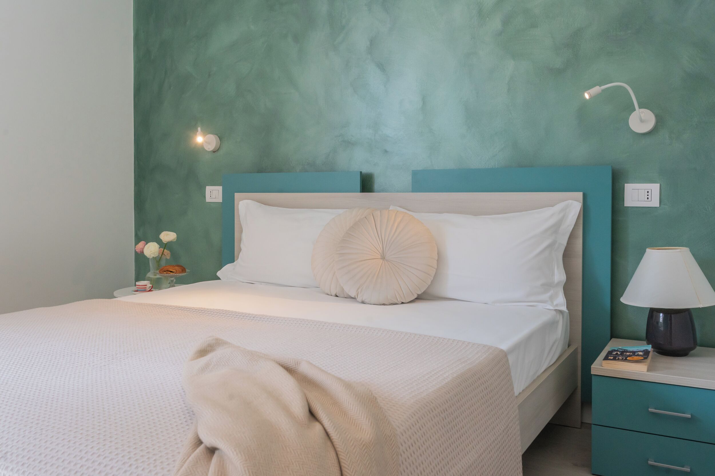 A neatly made bed with a white comforter and a beige throw blanket, a teal nightstand with a lamp, and a wall-mounted light fixture.