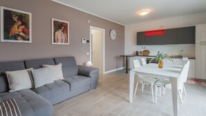 Un salon moderne avec un canapé gris, une table à manger avec des chaises blanches et un coin cuisine doté de meubles gris et d’une étagère rouge.