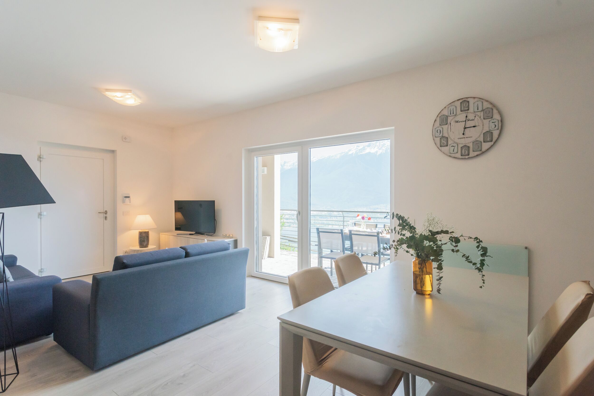 A modern living room with a sofa, a dining table with chairs, a TV, and a large window with a view.