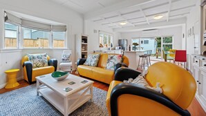 Amplia sala de estar con un sofá amarillo, una mesa de centro blanca y una mesita auxiliar amarilla. Se ve un área de cocina al fondo, con una silla roja y un armario blanco.