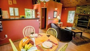 A cozy dining area with a wooden table set for two, surrounded by chairs. A stone fireplace and a small table with a lamp are visible in the background.