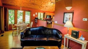 A cozy cabin interior with a wooden ceiling, a black leather sofa, a dining table, and a kitchen area.