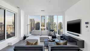Ein modernes Wohnzimmer mit großem Fernseher, grauen Sofas, einem Couchtisch und Blick auf die Stadt.