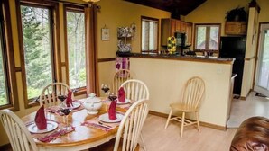 A dining area with a wooden table set for four, surrounded by white chairs. A kitchen with wooden cabinets and a black refrigerator is visible in the background.