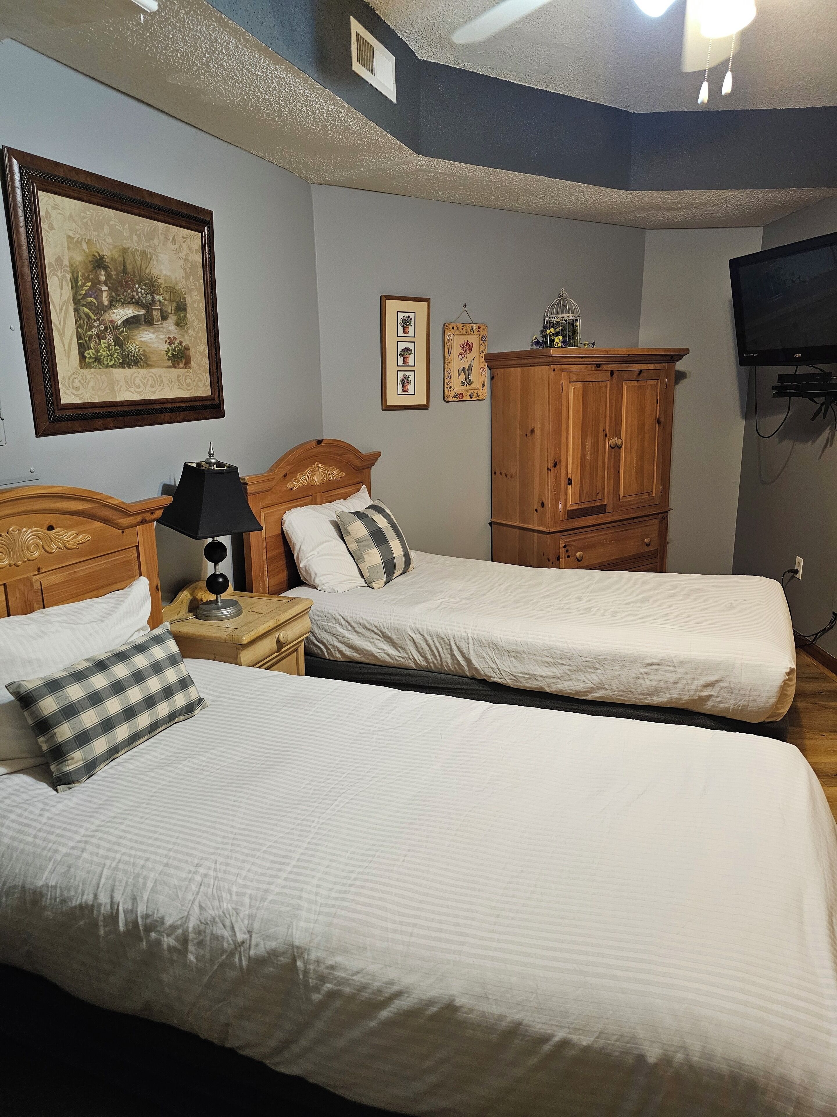 A bedroom with two single beds, a wooden wardrobe, a nightstand with a lamp, and a framed picture on the wall.