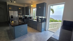 A modern kitchen with dark cabinets, a central island, and a view of the ocean through a sliding glass door. - 5 Bedroom, luxury private villa with pool & 360' ocean views (Gros Islet)