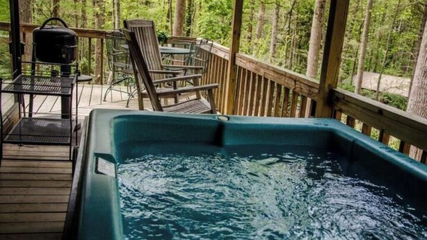Un jacuzzi en una terraza de madera con sillas y una mesa, rodeado de árboles.