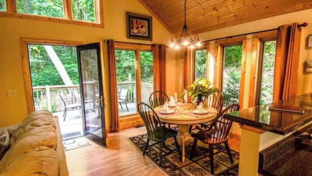 A cozy room with a wooden ceiling, a dining table set for four, and a large window overlooking a forested area. - Perfect Honeymoon cabin w/hot tub (Asheville)