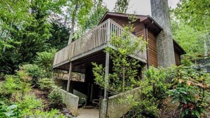 Une cabane dotée d’une terrasse en bois et d’une cheminée en pierre, entourée d’une forêt dense.