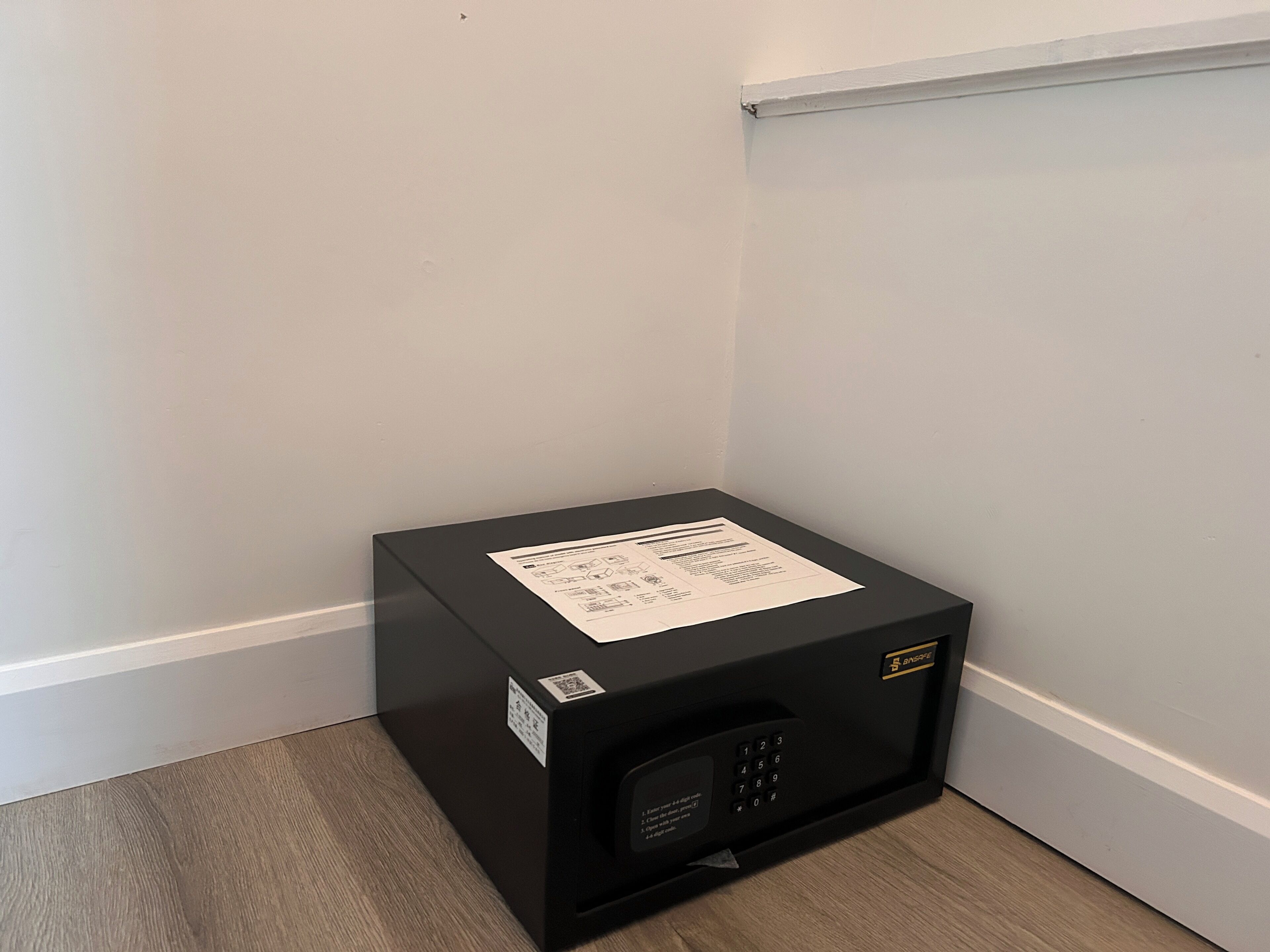 Een zwarte kluis met een QR-code en instructies aan de bovenkant, geplaatst op een houten vloer tegen een witte muur.