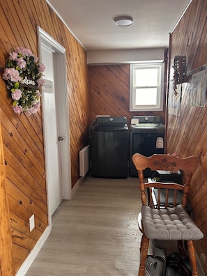 Un pasillo estrecho con paredes de madera y una puerta que dice “Bienvenido”. Una silla con cojín está apoyada contra la pared, y se ve un bote de basura al fondo.