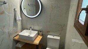 A compact bathroom with a white sink, a round mirror, a toilet, and a wooden shelf. - KYNTHIA seaview apartments (Samos)