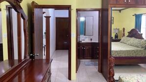 A room with a wooden wardrobe, a bed with a floral bedspread, and a bathroom with a sink and mirror. - 2-Bedroom/Bath Comfortable AI Residence- 
A/C, WiFi and 7 mins away from beach. (Statia Valley)