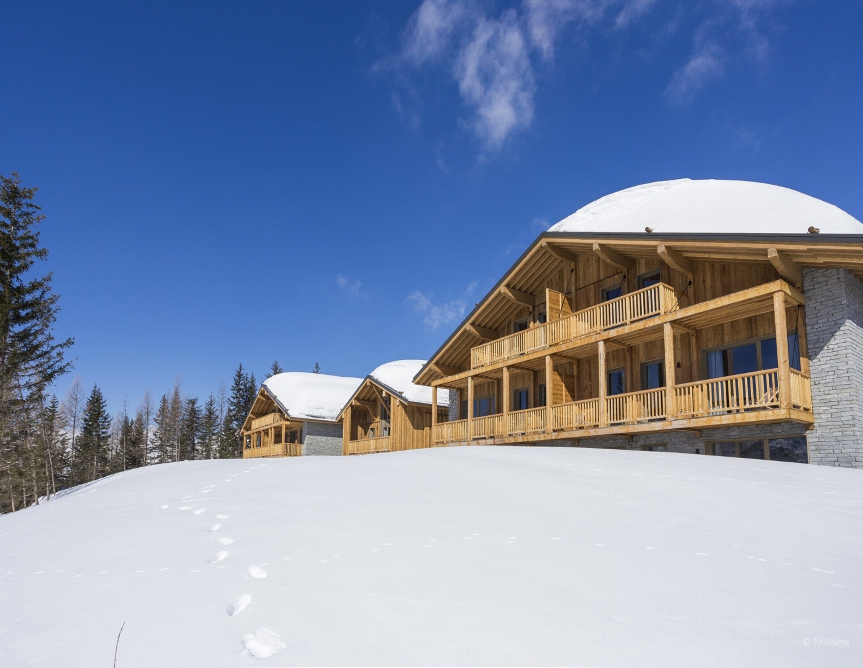 Un chalet en bois, avec un toit enneigé, des balcons en bois et un ciel d’un bleu limpide.