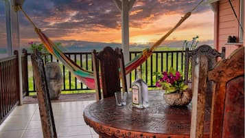 A wooden table with a bottle of liquor and a glass, set on a porch with a view of a sunset.