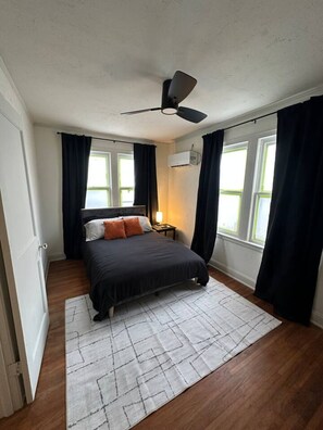 A bedroom with a bed, a ceiling fan, an air conditioning unit, and a window with curtains.