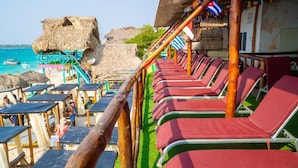 A beachside lounge area with rows of red lounge chairs, a thatched-roof structure, and a view of the sea.