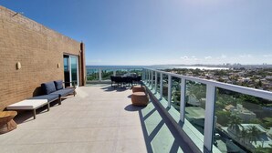 A rooftop terrace with a brick wall, a glass railing, a sofa, a table, and a view of the city and ocean. - COPDS0108 - Pedra do Sal Privilege (Salvador)