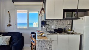 A compact living space with a kitchen, a sofa with cushions, a dining area, and a view of the sea. - COPDS0108 - Pedra do Sal Privilege (Salvador)