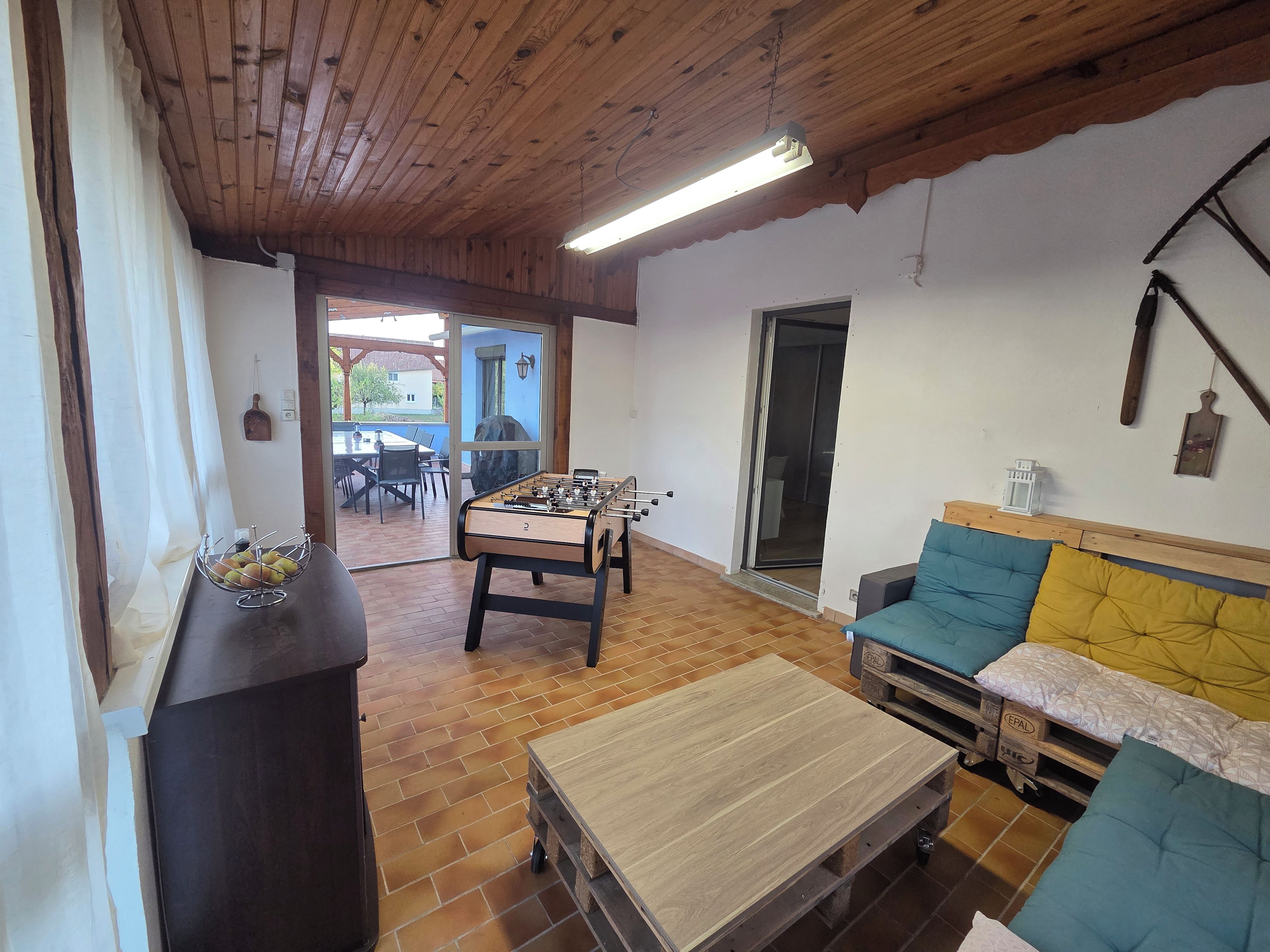 Une pièce avec un plafond en bois, une table de baby-foot, un canapé avec des coussins, une table sur laquelle se trouve un panier de fruits, et une porte-fenêtre coulissante donnant sur un espace extérieur.