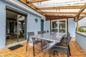 Eine überdachte Terrasse mit einem Glastisch und Stühlen, einer Holzbeschichtung und einer Glasschiebetür, die zu einem Innenbereich führt.