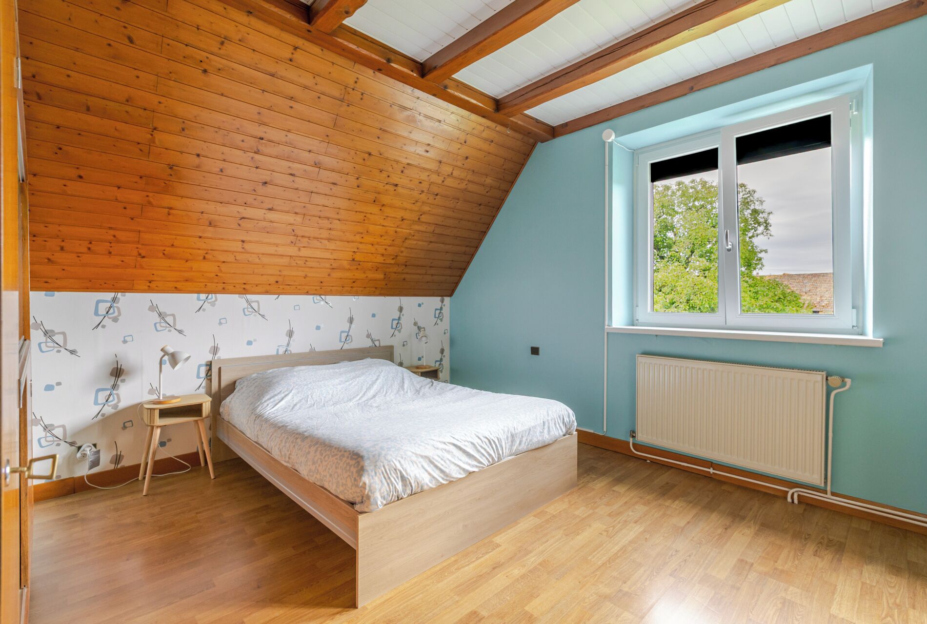 Un dormitorio con techo de madera, una cama, una mesita de noche, un ventanal con vistas y un radiador.