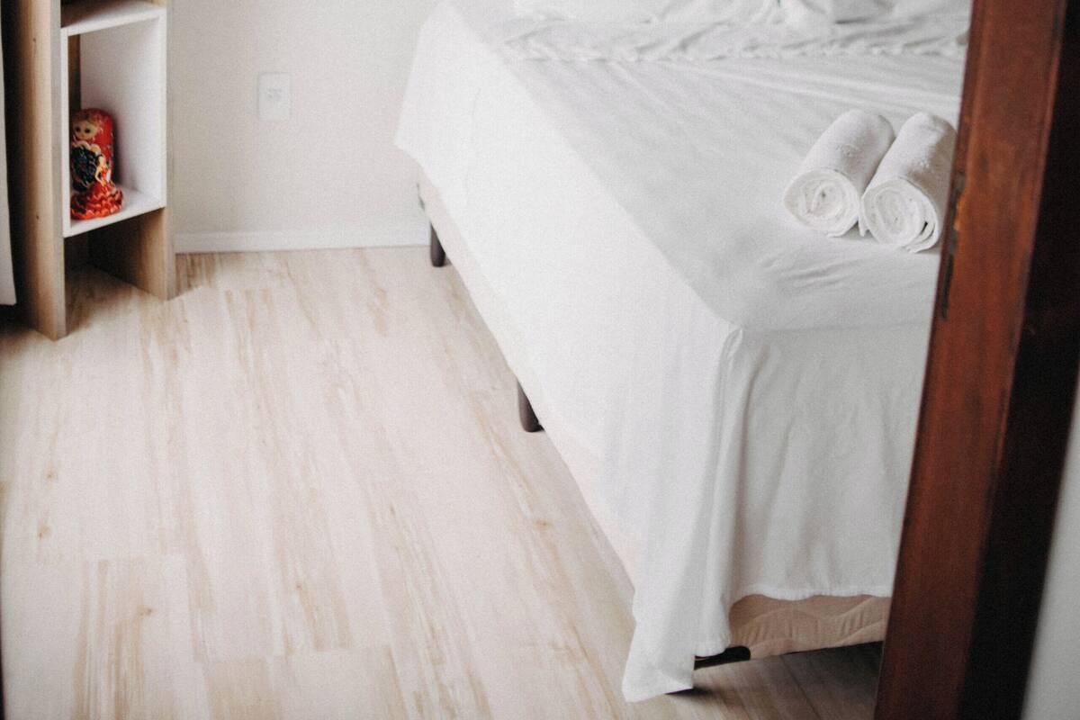 A bedroom with a bed, white linens, a wooden shelf, and an air conditioning unit.
