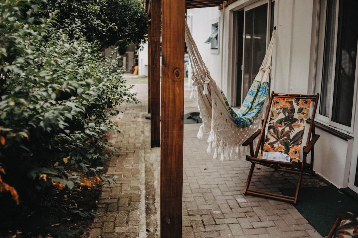 A hammock hanging between wooden posts, a wooden easel with a painting, and a brick pathway leading to a building.