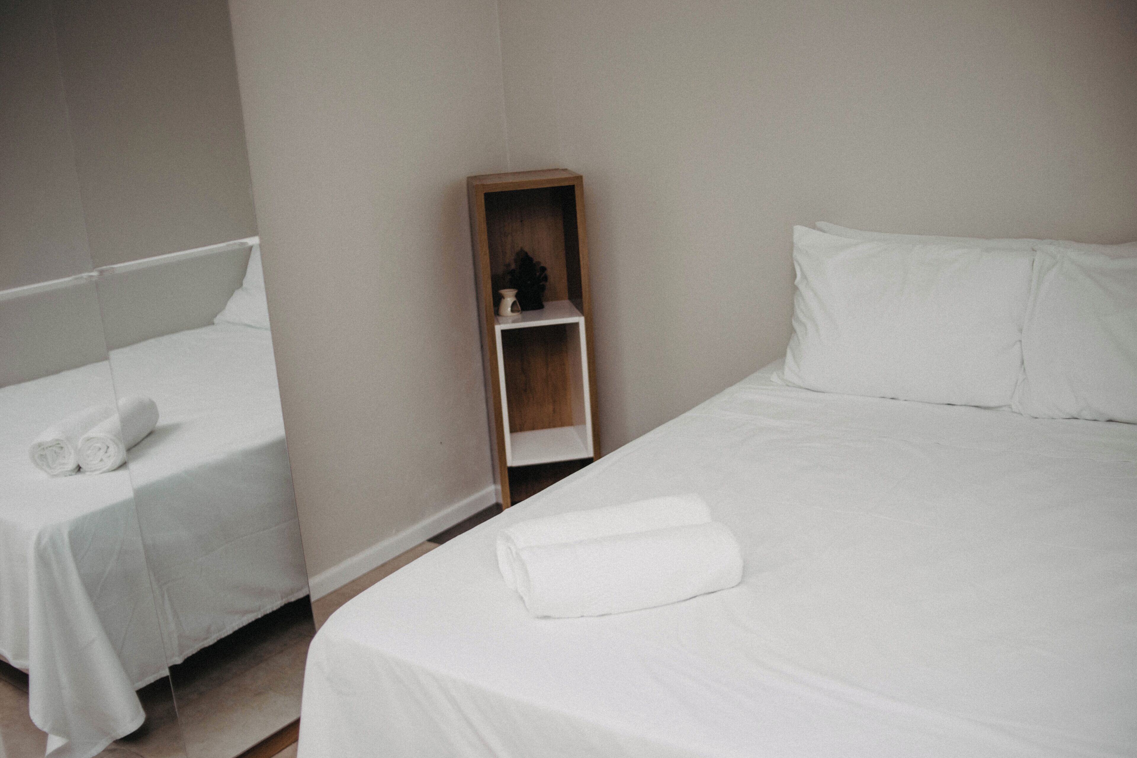 A hotel room with two beds, each with white linens and pillows, and a small wooden shelf with a vase and a book.