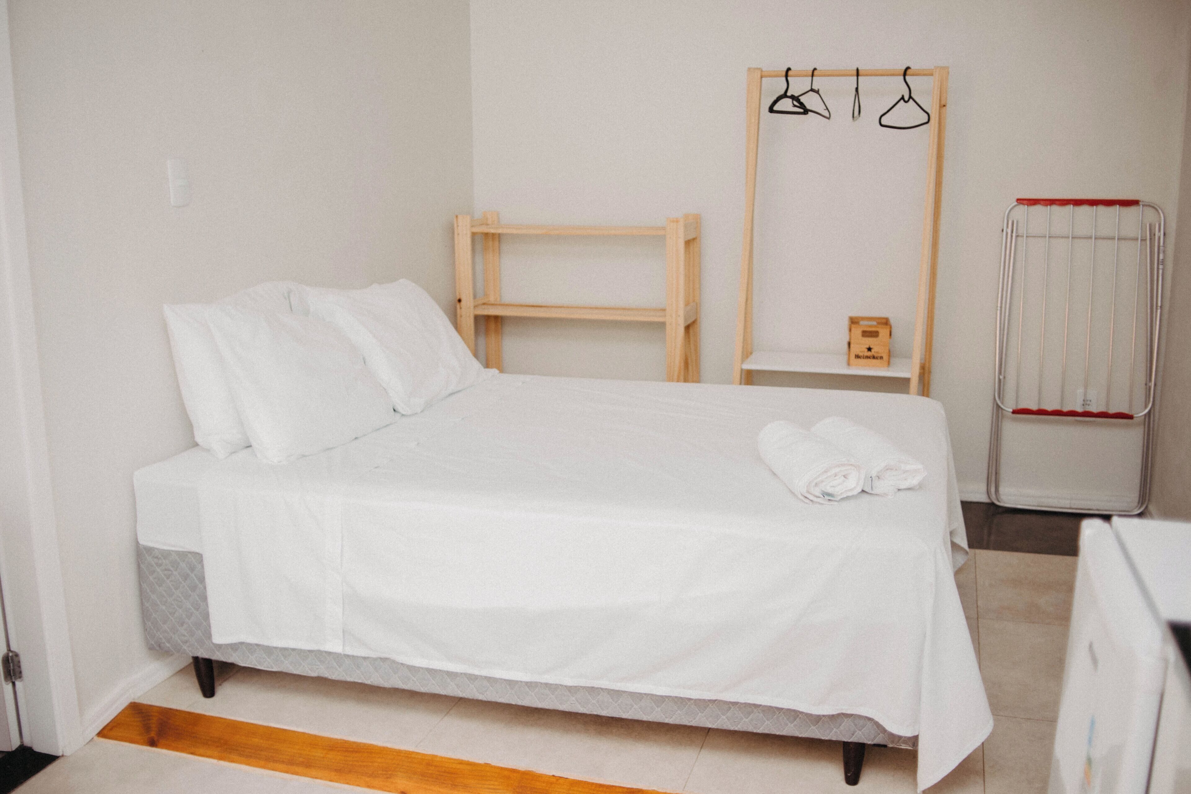 Un lit bien fait, avec du linge de lit blanc, une table de chevet en bois avec un compartiment, un porte-vêtements suspendu et un étendoir métallique.