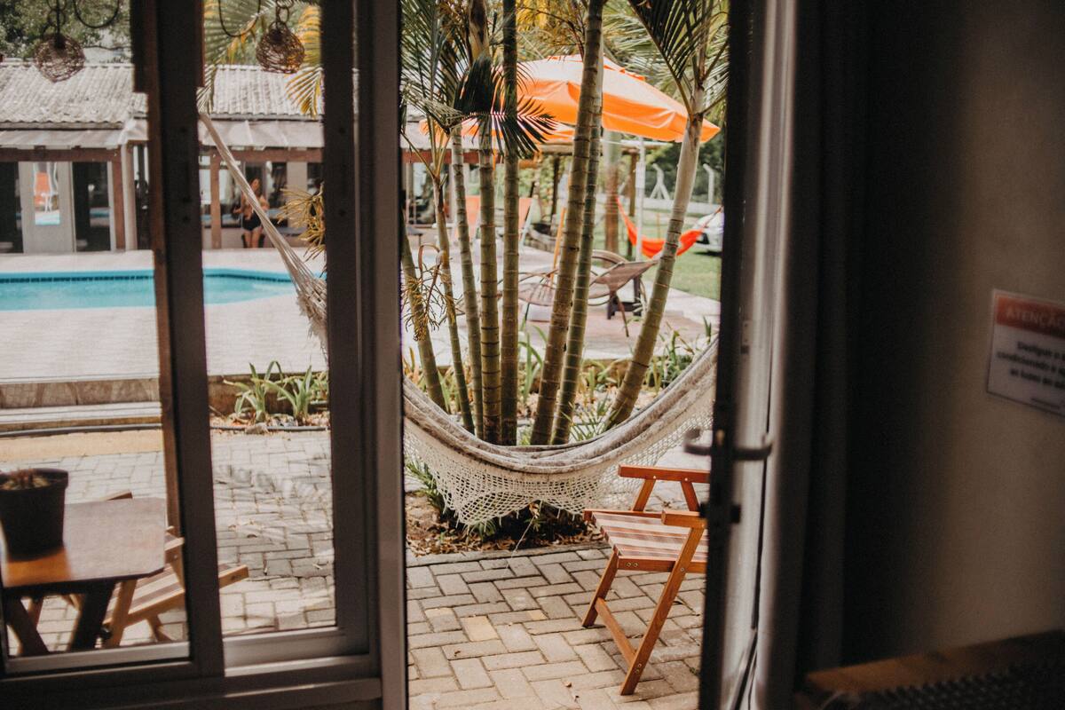 A view through a glass door of a poolside area with a hammock, wooden chairs, and an orange umbrella.
