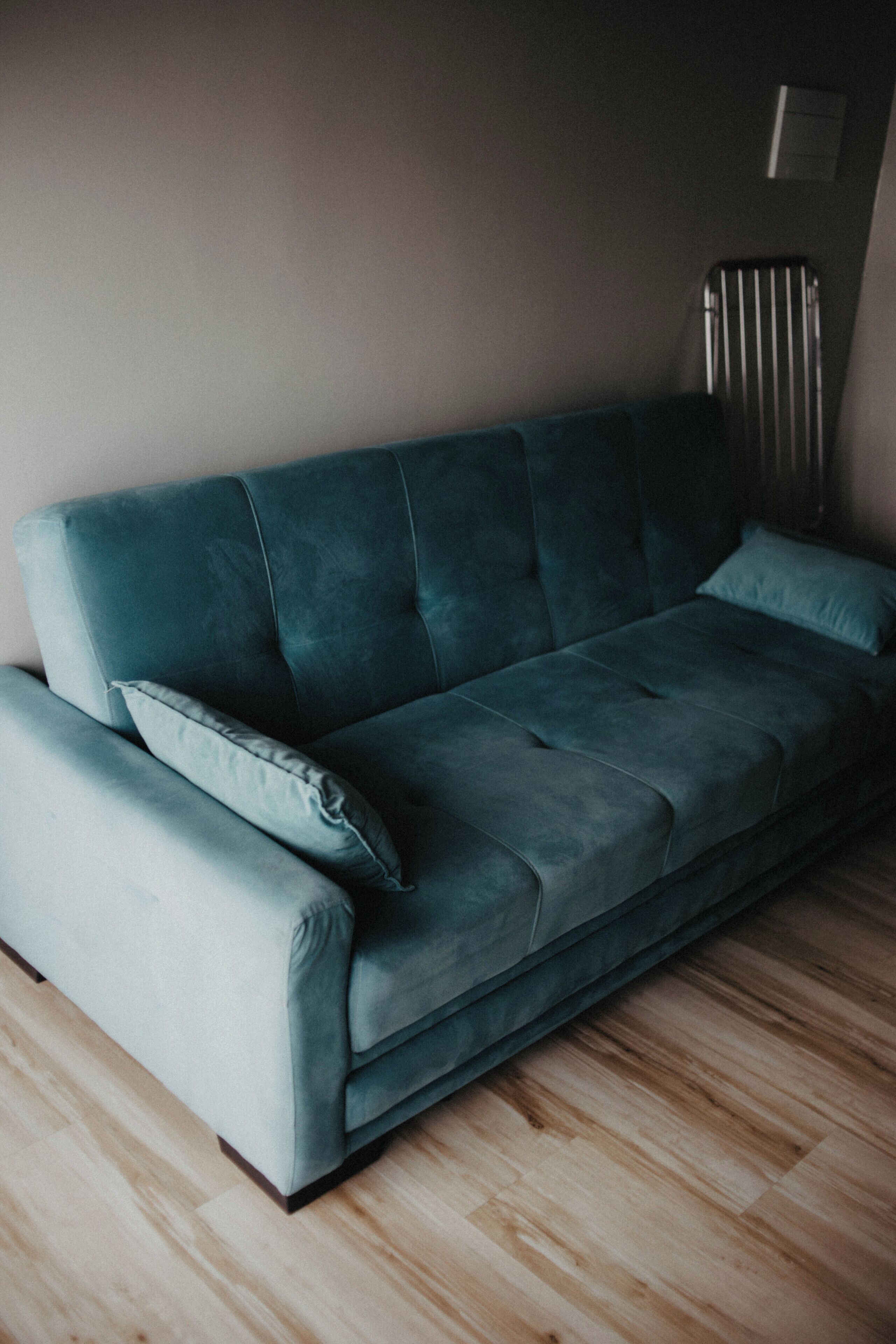 A teal sofa with a matching pillow in a room with wooden flooring and a radiator on the wall.