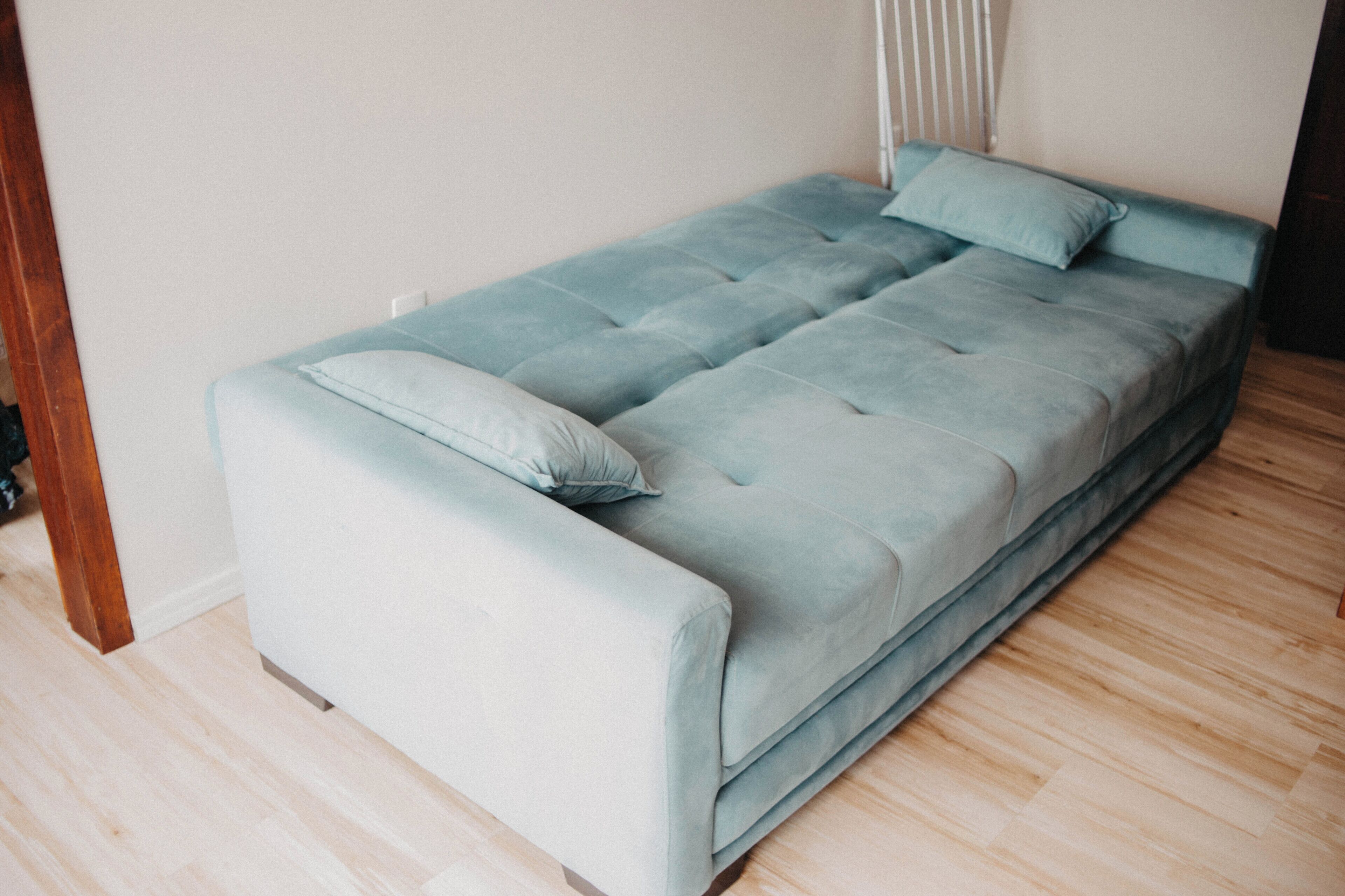 Un divano letto azzurro chiaro con cuscino coordinato, appoggiato a una parete bianca con pavimento in legno.
