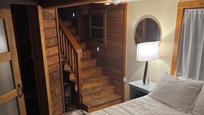 A bedroom with a bed, a nightstand, a lamp, a window with curtains, a wooden staircase, and a door leading to another room.