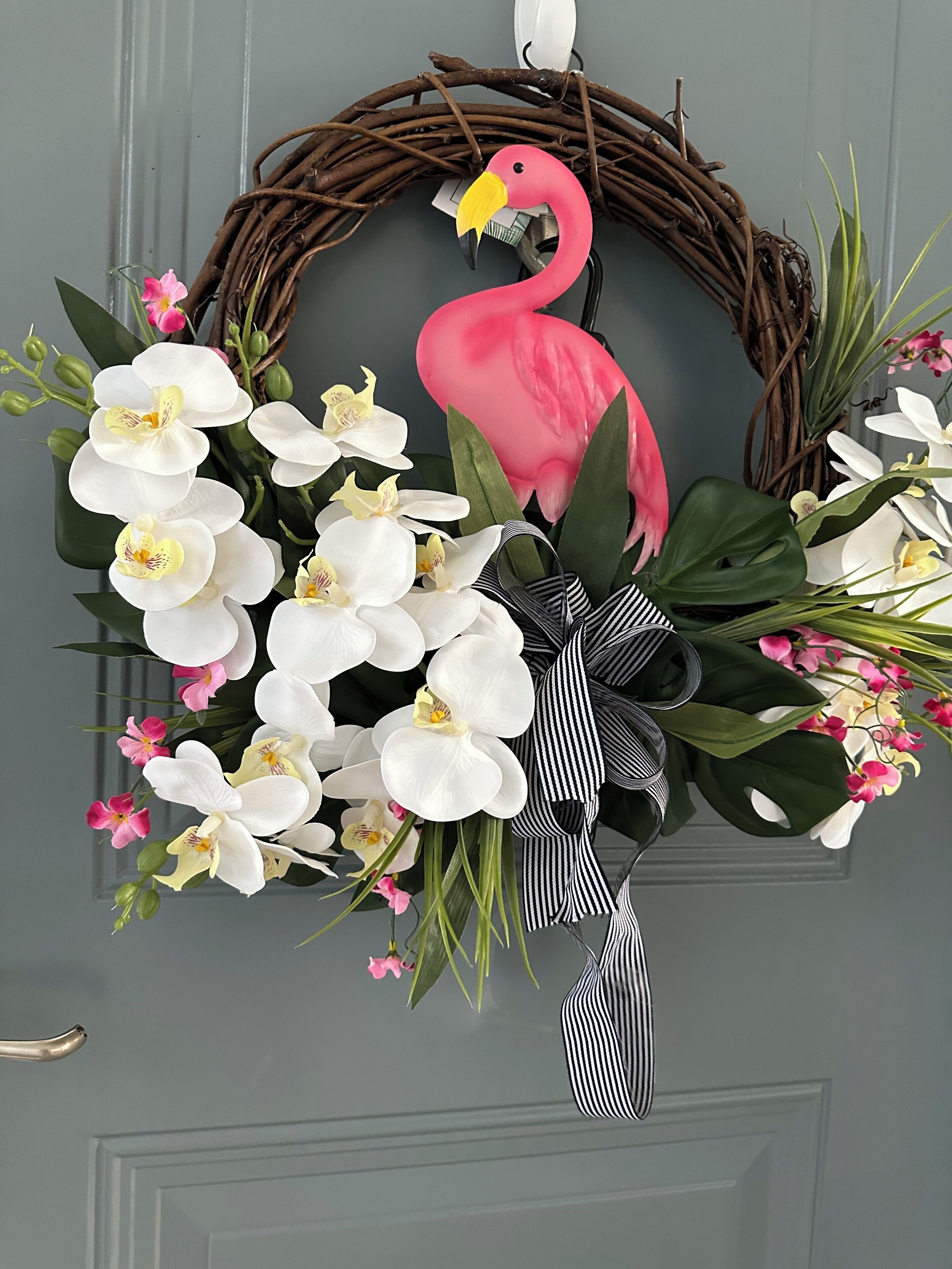A wreath with a flamingo decoration, white and pink orchids, and green foliage on a gray door.