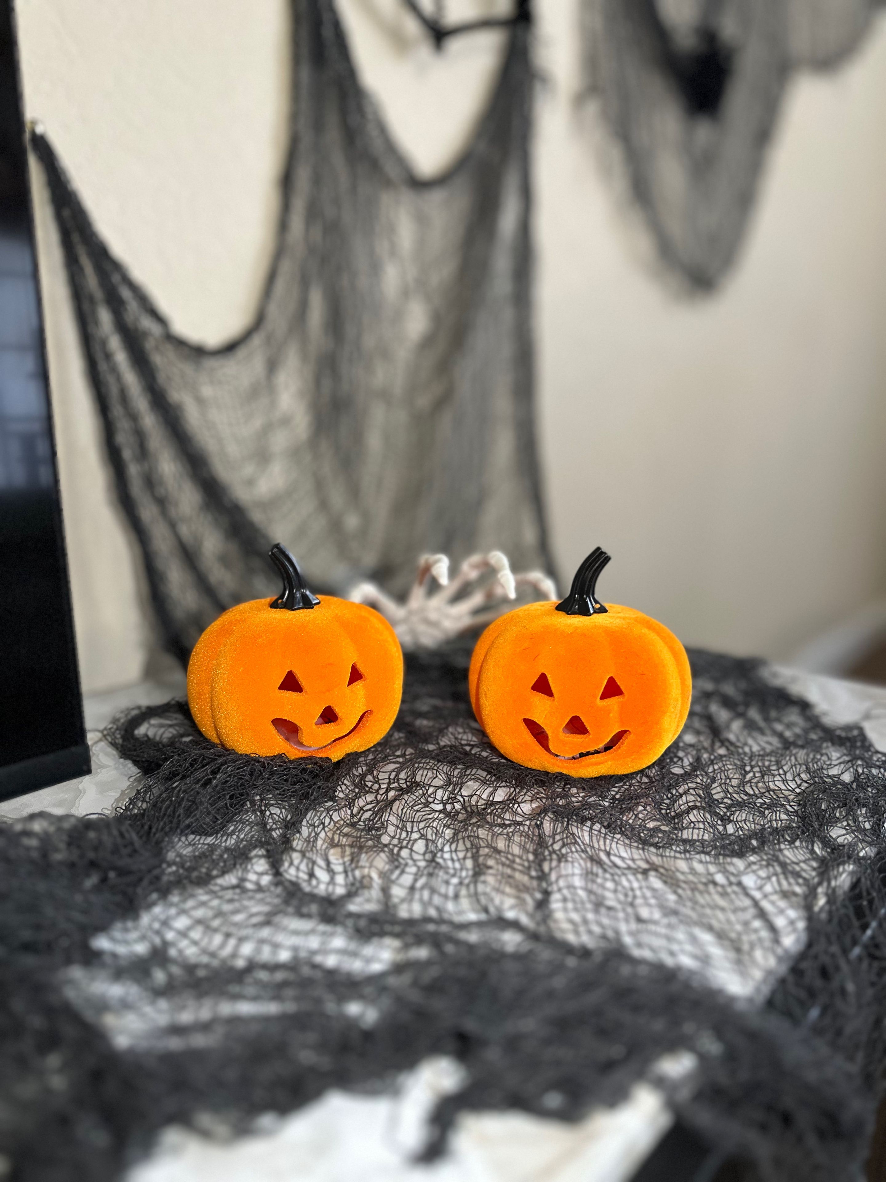 Deux petites citrouilles orange, décorées de visages, sont posées sur une table recouverte d’une nappe noire en dentelle.