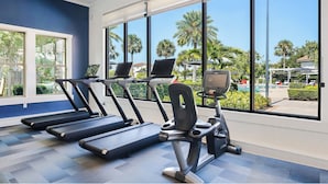 Un gimnasio con cintas de correr y equipo de ejercicio, ventanas amplias con vista a palmeras y una piscina.