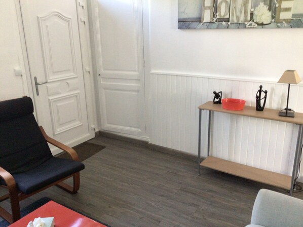 A room with a wooden floor, a black chair, a red table, and a small table with a lamp and decorative items. - Sara's home 24. (Rouen)