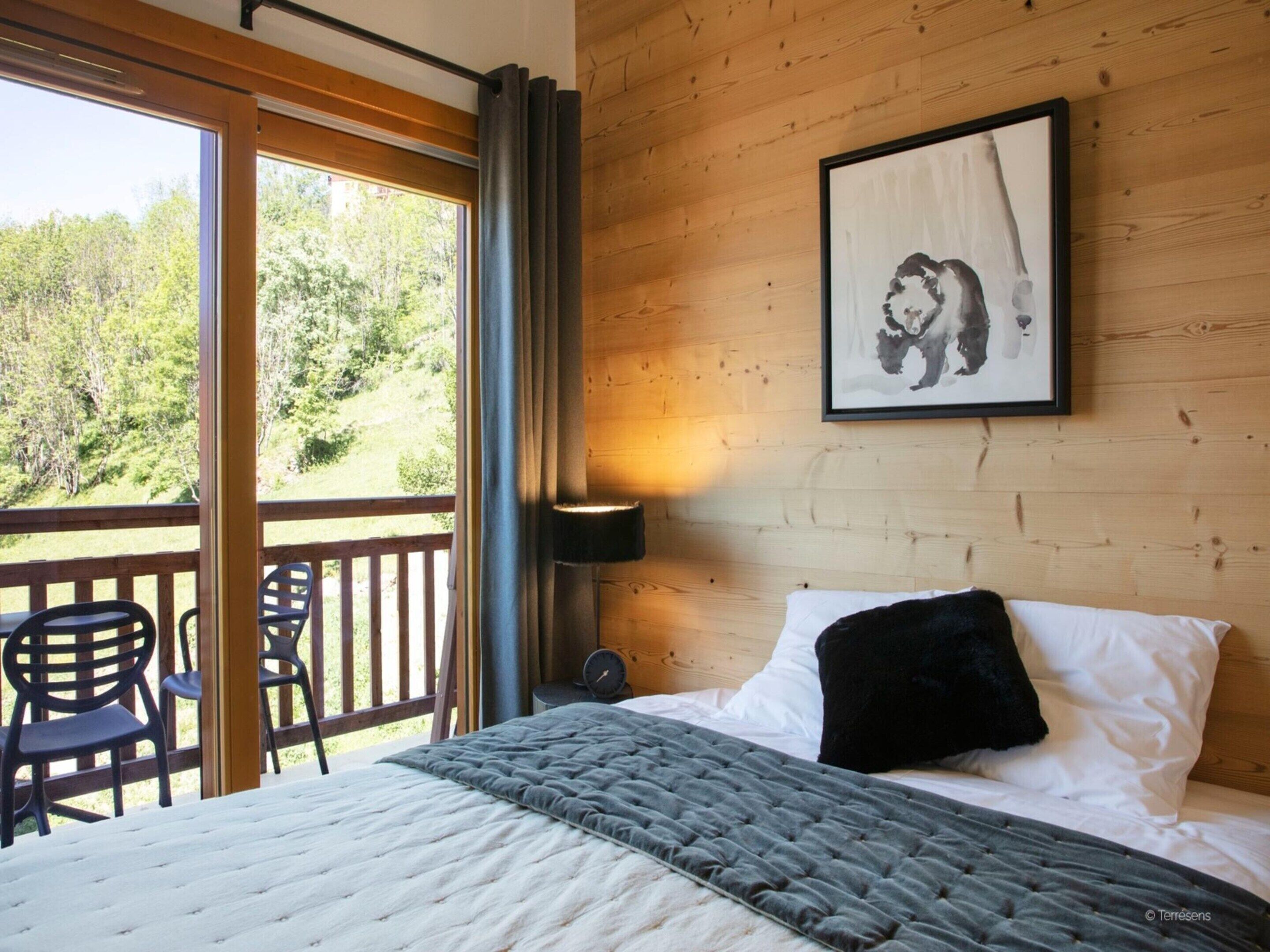 Un dormitorio con cama, ventana con cortinas, pared de madera, un cuadro enmarcado y vista a árboles.