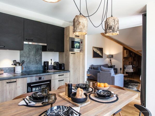 A modern kitchen with dark cabinets, a wooden dining table set with plates, glasses, and a bowl of fruit. - Chalet in Vaujany With Panoramic Mountain Views (Vaujany)