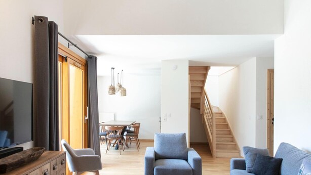 A modern living room with a sofa, armchairs, a coffee table, and a TV. There's a dining area with a table and chairs, and a staircase leading upstairs. - Chalet in Vaujany With Panoramic Mountain Views (Vaujany)