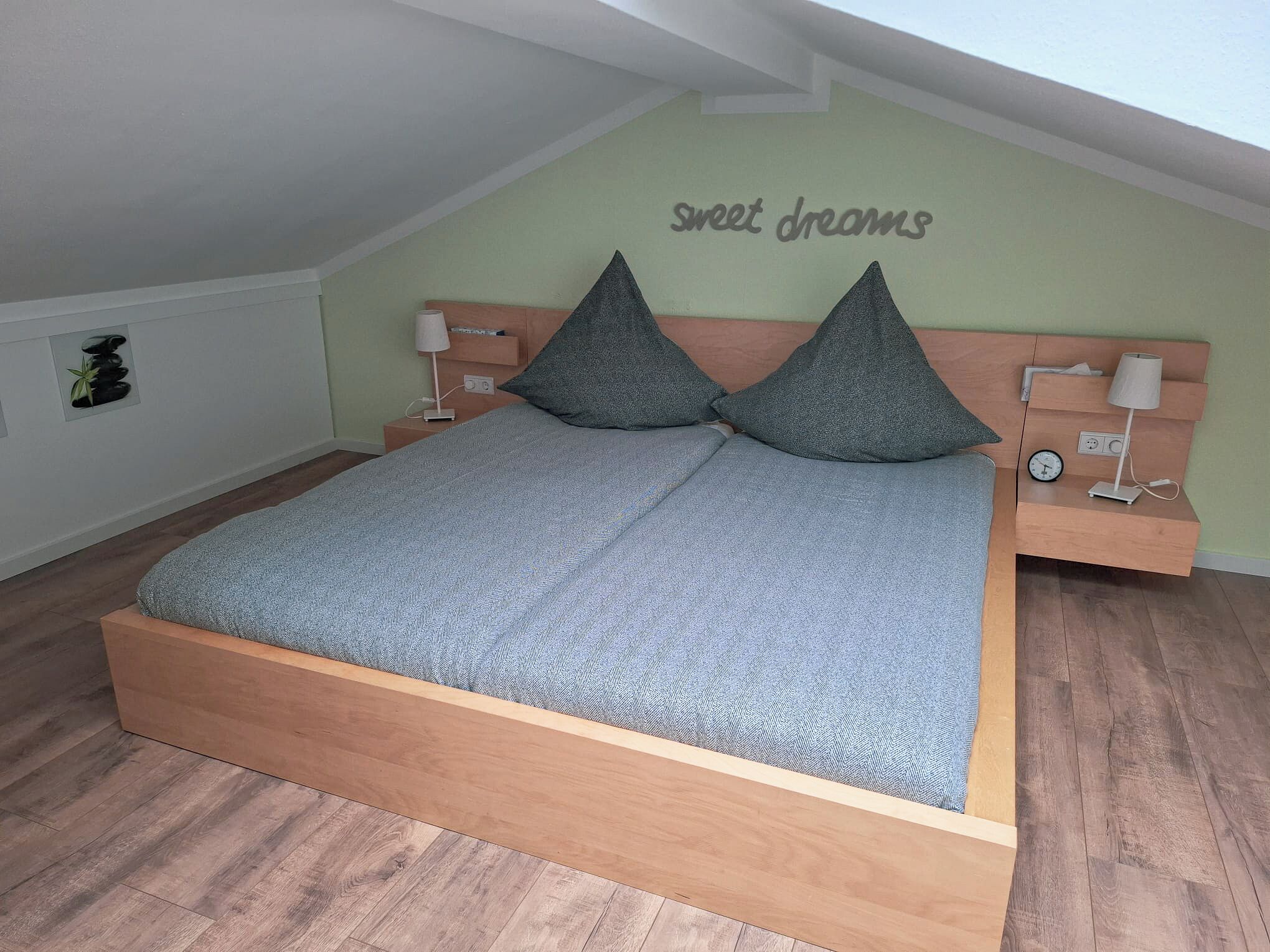 Een slaapkamer met een houten bed, twee kussens, een nachtkastje met een lamp, en een muur waarop staat: "sweet dreams".