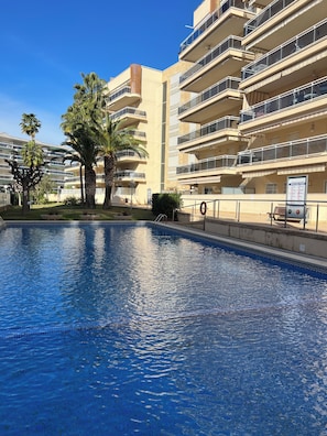 A swimming pool with a clear blue water surface, surrounded by a building with multiple balconies and palm trees.