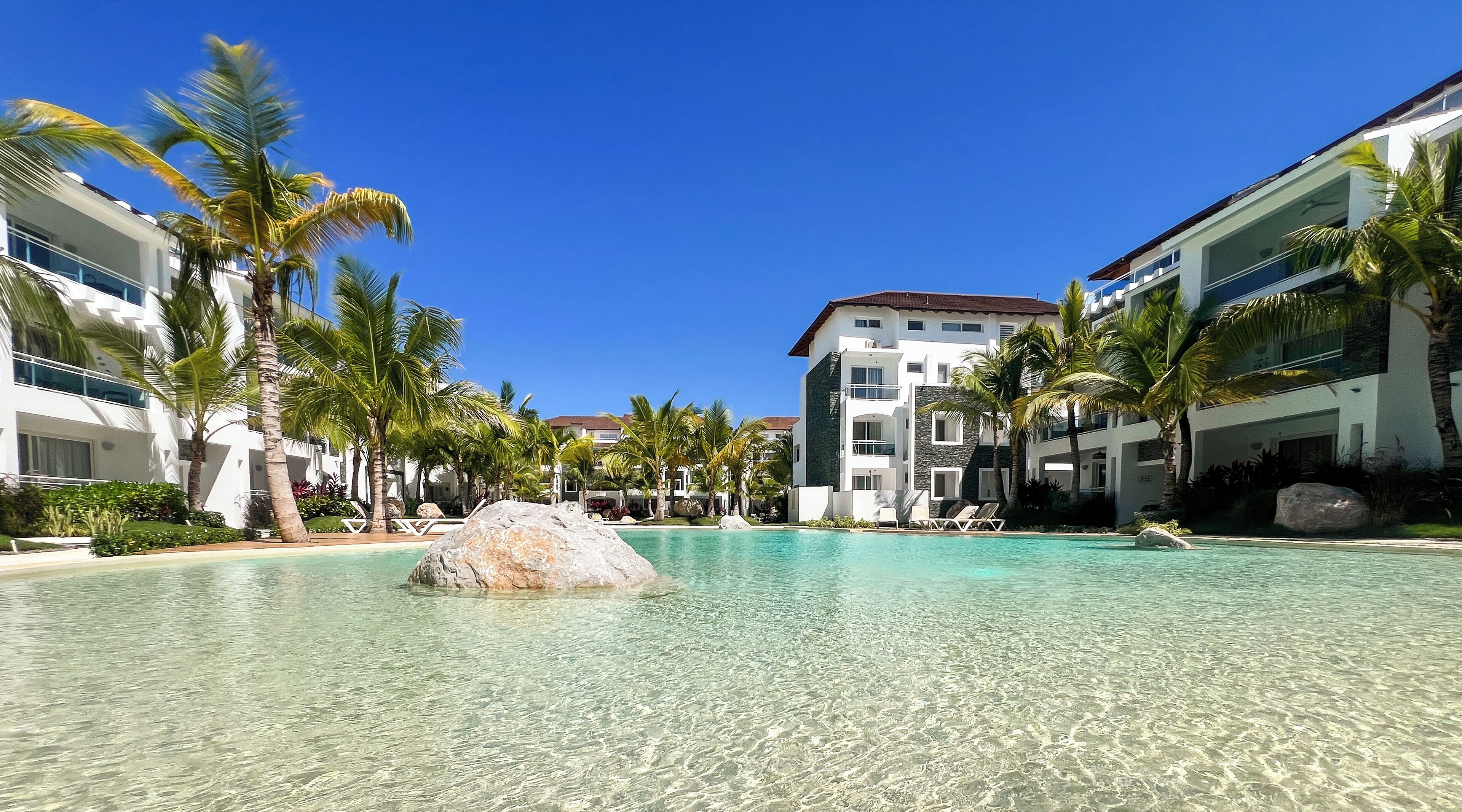 A resort with a clear pool, palm trees, and modern buildings.